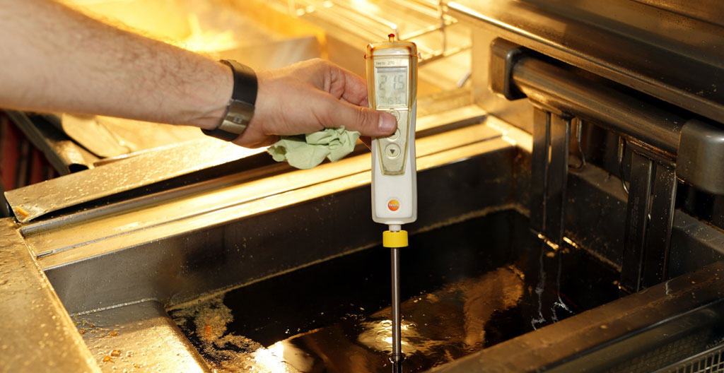 Technicien collecteur accroupi dans une cuisine professionnelle, mesurant un espace de stockage avec son matériel d'évaluation, équipements inox en arrière-plan